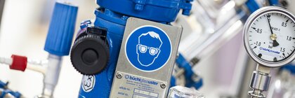 Eine Laborausrüstung mit einer blauen Vorrichtung, einem Druckmesser und verschiedenen Ventilen. Ein Warnschild für Augenschutz ist sichtbar.