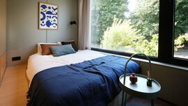 Gemütliches Schlafzimmer mit großem Fenster, blauer Decke auf dem Bett, abstrakter Kunst an der Wand, rundem Tisch und Bäumen draußen.