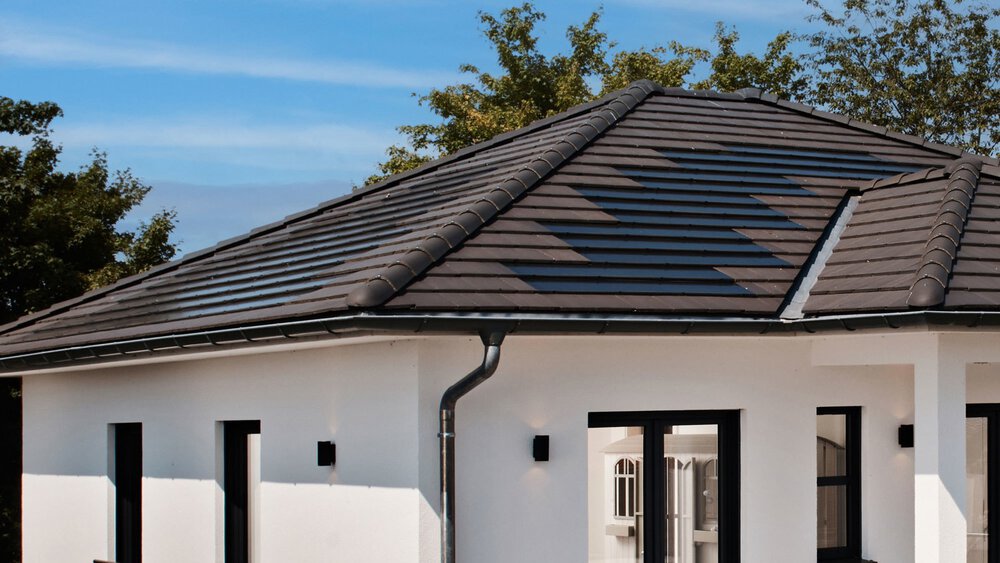 Ein modernes weißes Haus mit dunklem Ziegelwalmdach und großen Fenstern vor einem blauen Himmel und Bäumen.