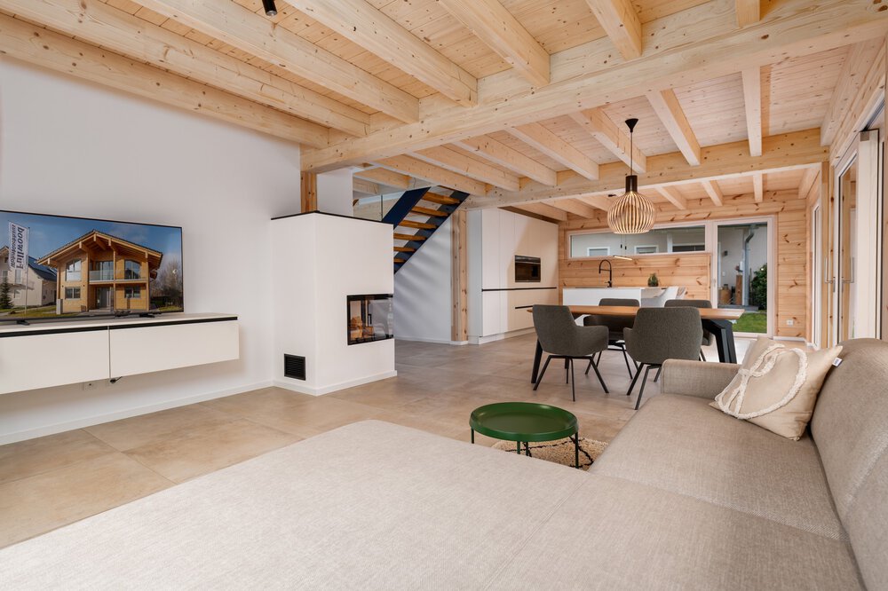 Modernes offenes Wohnzimmer mit Holzbalken, grauem Sofa, Fernseher, Esstisch und Küche im Hintergrund.