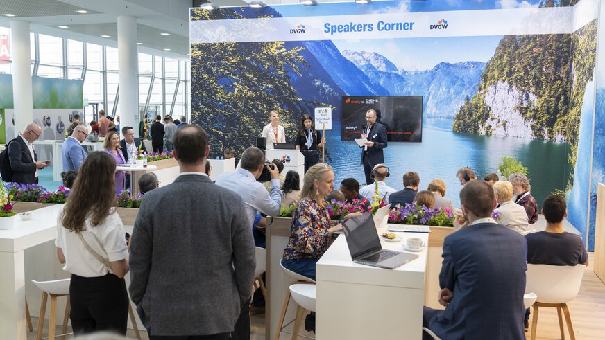 Belebte Messe-Ecke 'Speakers Corner' mit Rednern. Das Publikum an Tischen mit Laptops, Getränken & Deko.