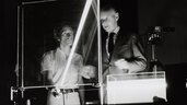 Black and white photo of two men behind an experimental setup where laser light is directed through a glass plate and a container filled with liquid.