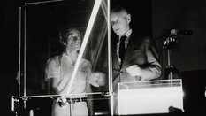 Schwarzweißfoto von zwei Männern hinter einem Versuchsaufbau bei dem Laserlicht durch eine Glasplatte und einen mit Flüßigkeit gefüllten Behälter gelenkt wird.