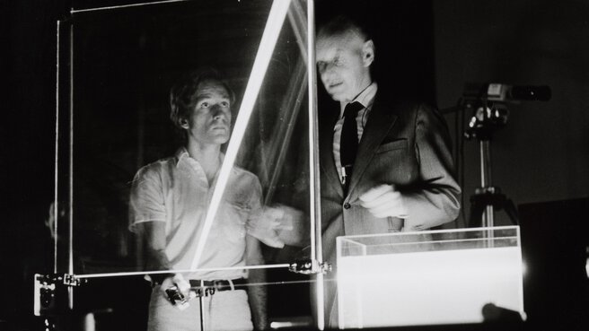 Black and white photo of two men behind an experimental setup where laser light is directed through a glass plate and a container filled with liquid.