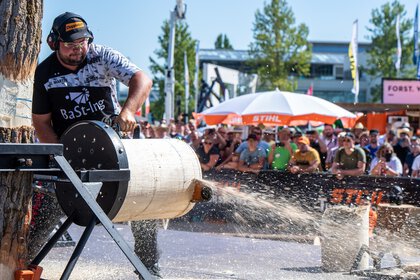 In a show area on the open-air grounds of INTERFORST, a man saws through a horizontally mounted tree trunk with a chainsaw in front of an audience.