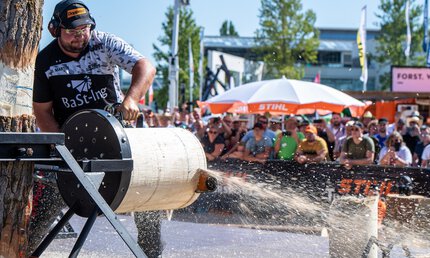 In a show area on the open-air grounds of INTERFORST, a man saws through a horizontally mounted tree trunk with a chainsaw in front of an audience.