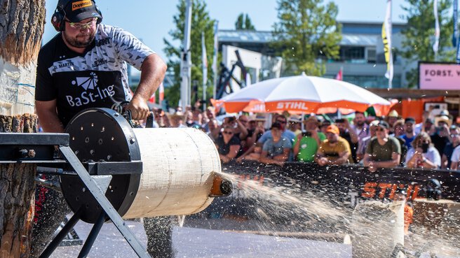 In a show area on the open-air grounds of INTERFORST, a man saws through a horizontally mounted tree trunk with a chainsaw in front of an audience.