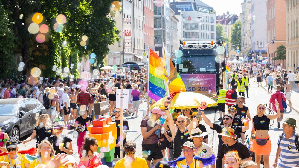 A colorful Pride parade with rainbow flags, soap bubbles, and a large crowd winding its way down a sunny city street.