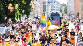 A colorful Pride parade with rainbow flags, soap bubbles, and a large crowd winding its way down a sunny city street.