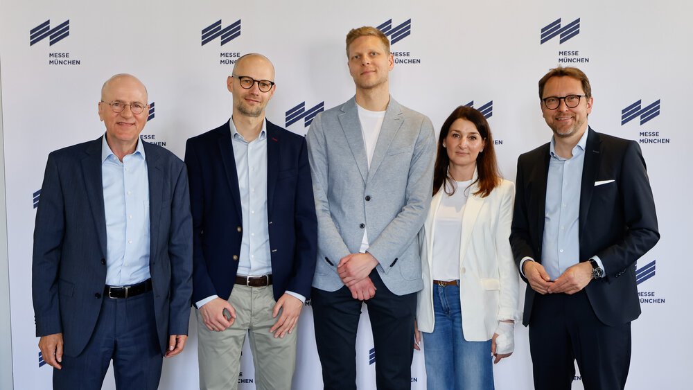 Four men and a woman are standing in front of a white press wall of Messe München, smiling and posing for a group photo.