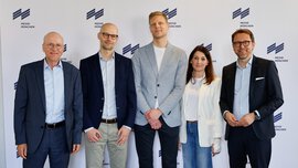 Four men and a woman are standing in front of a white press wall of Messe München, smiling and posing for a group photo.