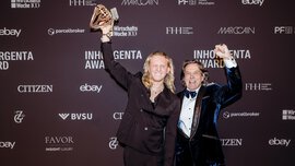 Two smiling men with long hair raise their arms in the air in front of a photo wall of the INHORGENTA AWARD and hold up a trophy.