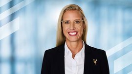  Smiling woman with long blonde hair, glasses, and business attire in front of a blue background.