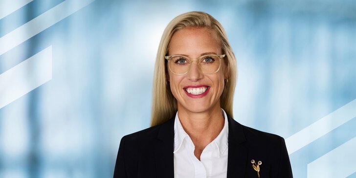  Smiling woman with long blonde hair, glasses, and business attire in front of a blue background.