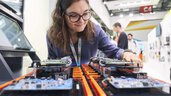 A woman with glasses examines electronic components on a circuit board on display at an exhibition stand.