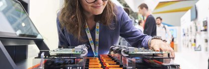 A woman with glasses examines electronic components on a circuit board on display at an exhibition stand.