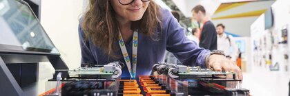 Eine Frau mit Brille untersucht elektronische Komponenten auf einer Platine, die an einem Messestand ausgestellt ist.