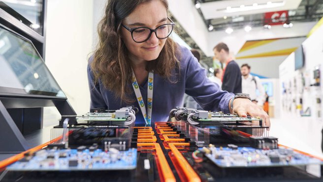 A woman with glasses examines electronic components on a circuit board on display at an exhibition stand.