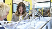 Two women smiling looking at a presentation in a brightly lit water tank at a trade fair stand.