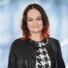 Eine lächelnde Frau mit schulterlangen dunklen Haaren, mit rötlichen Spitzen in einem schwarz-weiß gemusterten Oberteil und einer schwarzen Lederjacke.