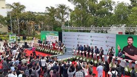 A crowd participates in an outdoor ribbon-cutting ceremony while officials stand on stage and photographers capture the event.