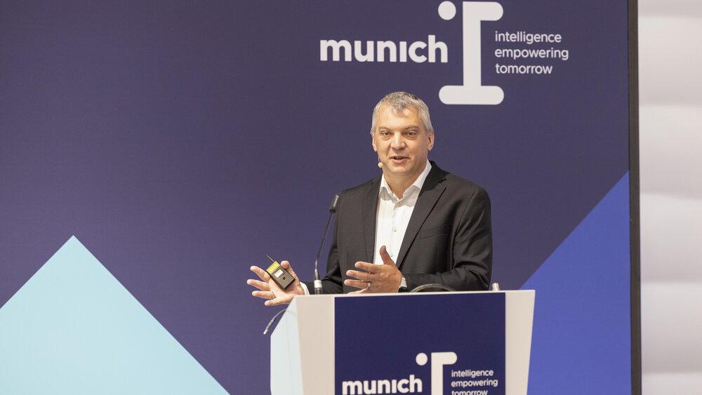 Ein Mann im Anzug und mit grauen Haaren hält an einem Podium der „munich i“ einen Vortrag.