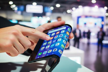 Auf dem Bild kann man einen Tablet Bildschirm sehen, der in der Mitte gefaltet ist, er wird von einer Männerhand auf einer Messe gehalten.