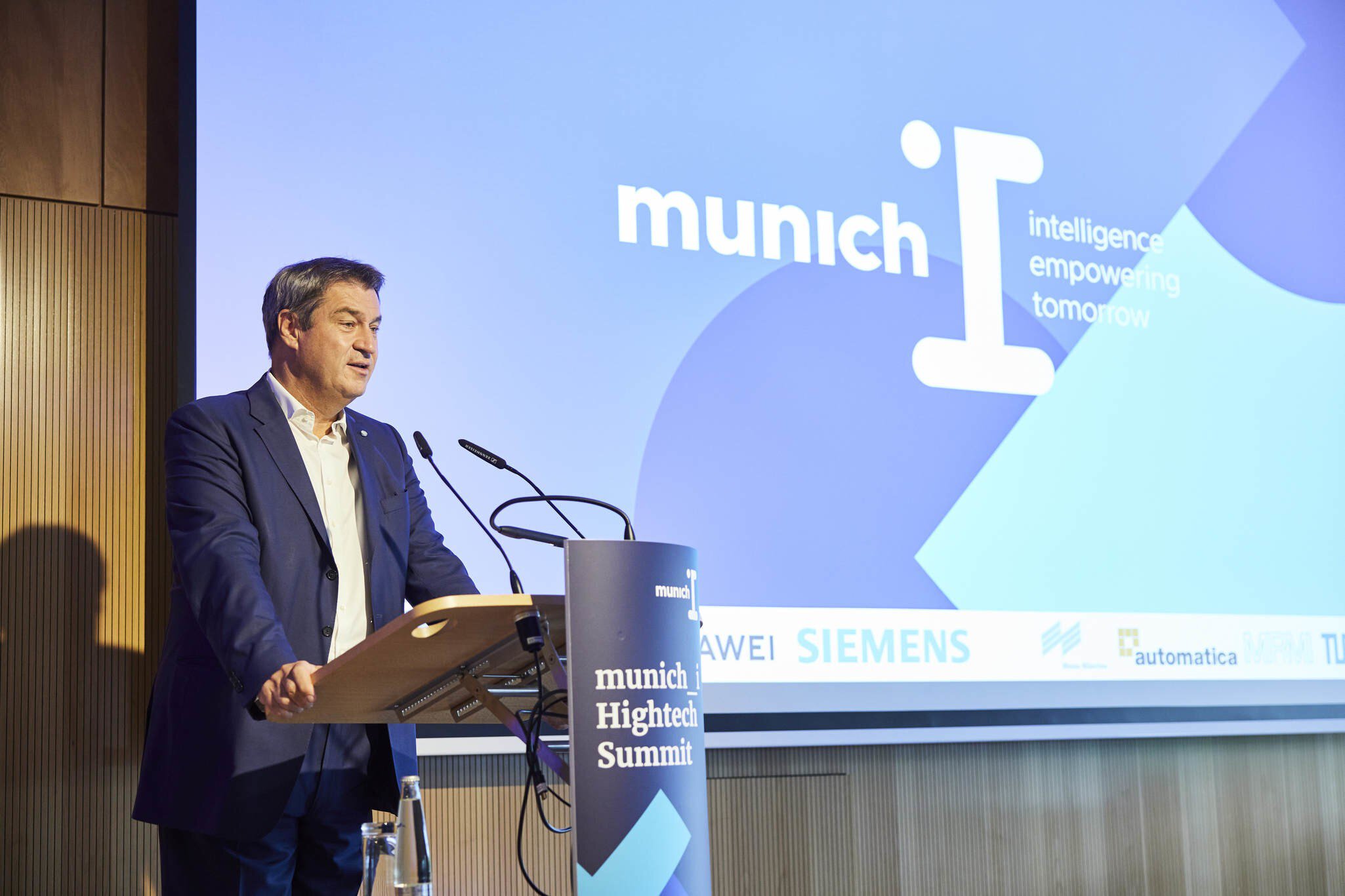 Ein Mann im blauen Anzug steht an einem Rednerpult zum munich-i Hightech Summit mit einer angestrahlten Leinwand im Hintergrund.