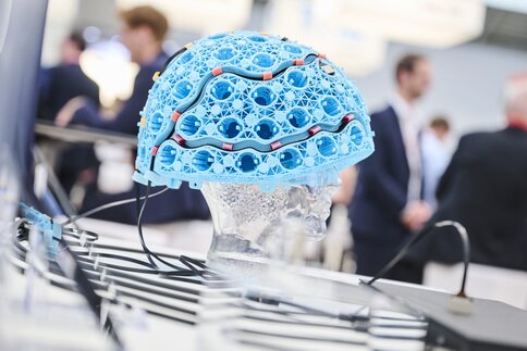 Durchsichtige Kopfattrappe mit einem blauen, gitterförmigen Helm, in den Kabel und Halterungen für optische Sensoren integriert sind, genutzt zur lichtbasierten Hirndiagnostik in der Biophotonik.