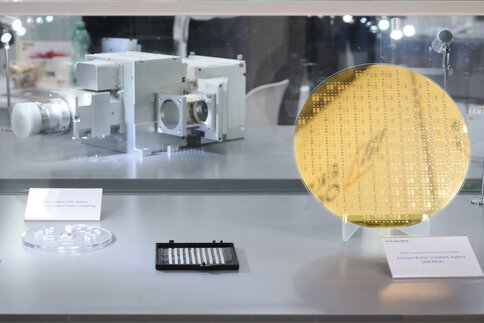 High-tech components from integrated photonics on display in a glass showcase. A large gold-colored wafer plate takes center stage.
