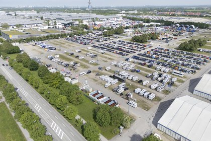 Luftaufnahme des großen Parkplatzes voller Autos und Wohnmobile vor dem Messeeingang Ost. Im Hintergrund sind die Messehallen zu sehen.