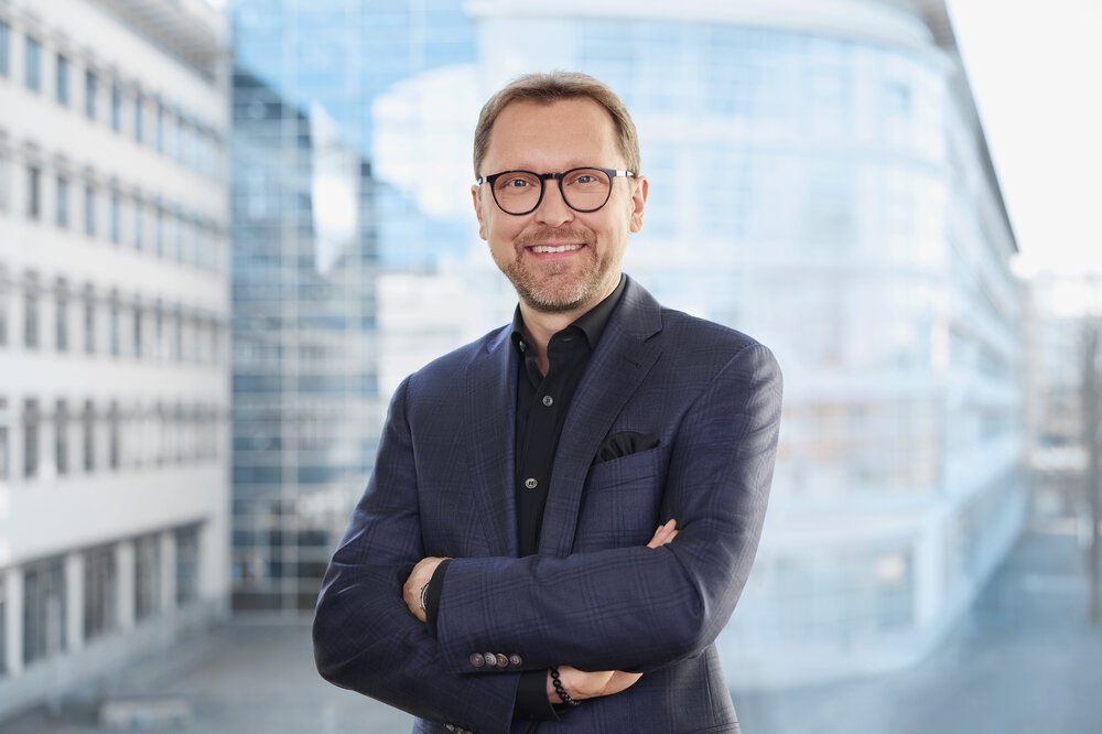 Lächelnder Mann in einem blaukarierten Anzug mit verschränkten Armen, der vor einem modernen Glasgebäude steht.