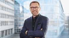 Lächelnder Mann in einem blaukarierten Anzug mit verschränkten Armen, der vor einem modernen Glasgebäude steht.