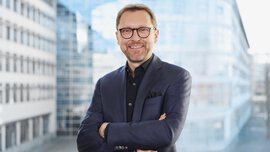 Lächelnder Mann in einem blaukarierten Anzug mit verschränkten Armen, der vor einem modernen Glasgebäude steht.