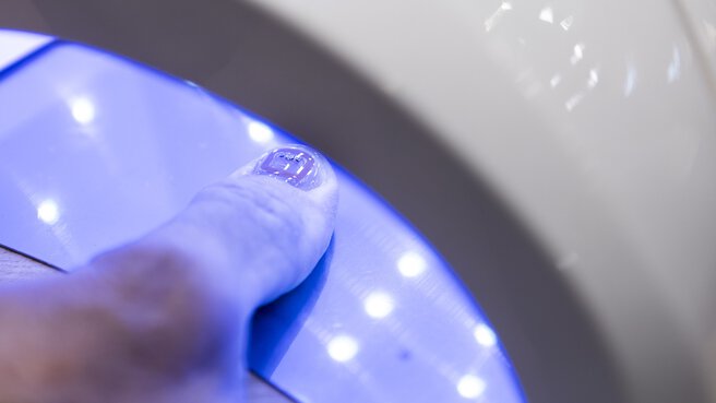 Close-up of a thumb under a UV lamp curing a glued nail with integrated RFID circuits.
