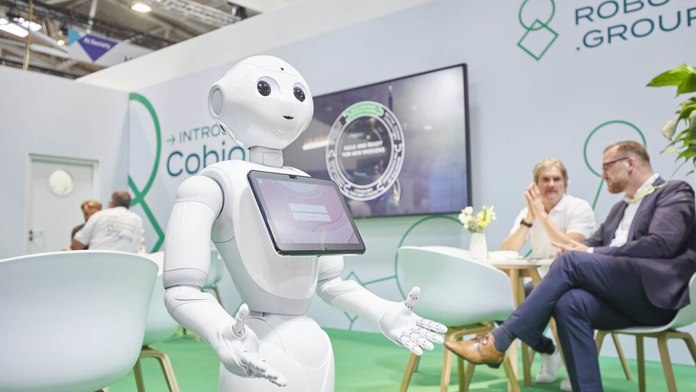 A white humanoid robot with a screen interacts at a technical exhibition stand while people sit in the background.