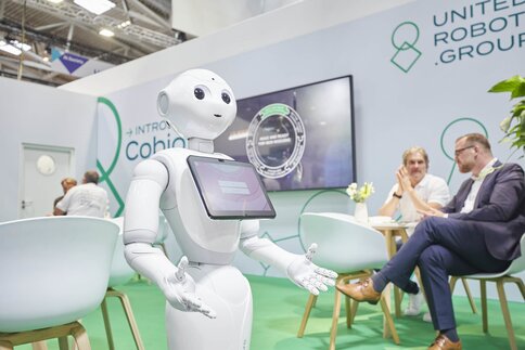A white humanoid robot with a screen interacts at a technical exhibition stand while people sit in the background.