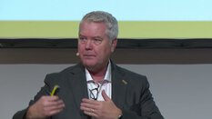 Ein Mann im Anzug spricht auf der Bühne und hält einen Clicker, trägt ein Headset-Mikrofon.