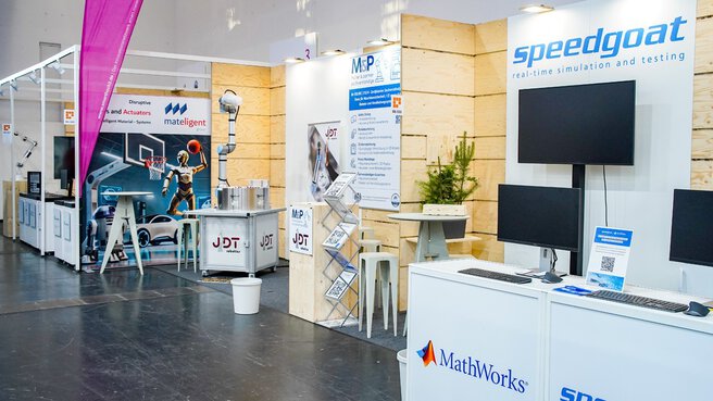 Trade fair booth with monitors, info counters and wooden panels, featuring Speedgoat and MathWorks logos in an exhibition hall.