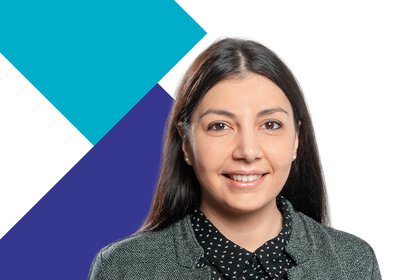 Lächelnde Frau mit langem, dunklem Haar trägt einen grauen Blazer und ein dunkles gepunktetes Hemd, im Hintergrund sind geometrische Formen zu sehen.