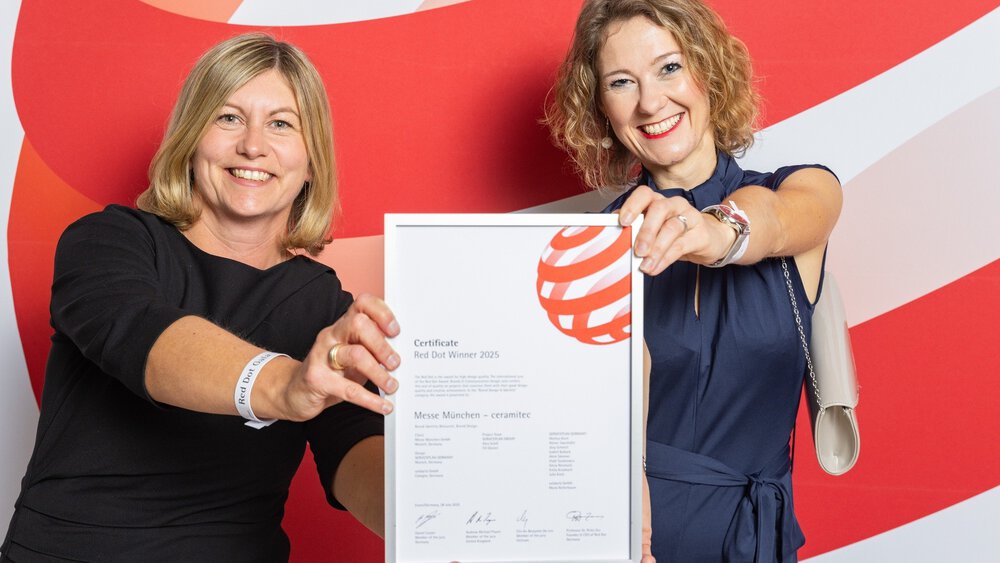 Two smiling women in formal attire hold up ceramitec's “Red Dot Winner 2025” certificate for the camera.