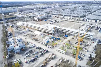 Luftaufnahme des Freigeländes der bauma 2025 mit mehreren Kränen, Containern und Fahrzeugen. Ein langes, mehrstöckiges Gebäude ist im Bau, umgeben von Materiallagern und Arbeitsbereichen. Im Hintergrund sind große Hallen und eine Stadtlandschaft zu sehen.