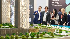 Mehrere Messebesucher betrachten ein großes Modell mit zwei Hochhäusern und Gebäuden mit einer Parkanlage und einem Strand daneben.