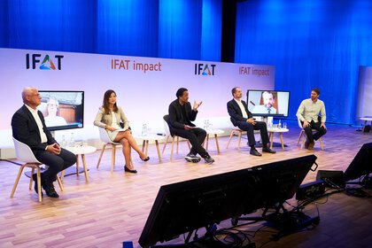 Podiumsdiskussion im Rahmen von „IFAT impact“ auf einer Bühne mit blauem Hintergrund; fünf Personen sitzen auf Stühlen, zwei zusätzliche Referentinnen oder Referenten werden auf Monitoren zugeschaltet, vor der Bühne stehen Kameras und Technik.