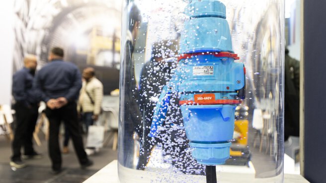 Blue industrial plug in a water cylinder with air bubbles; trade fair visitors talking in the background.