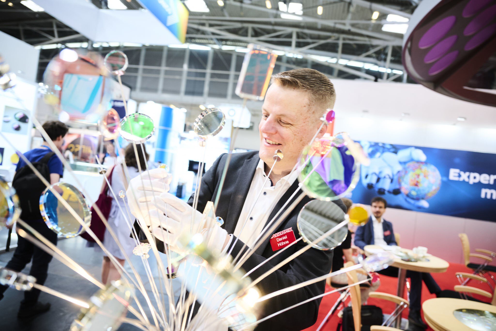 Ein lächelnder Mann im Anzug steht an einem Stand auf der Laser World of Photonics vor einem Strauß aus unterschiedlichen Linsen, die auf langen Antennen sitzen.