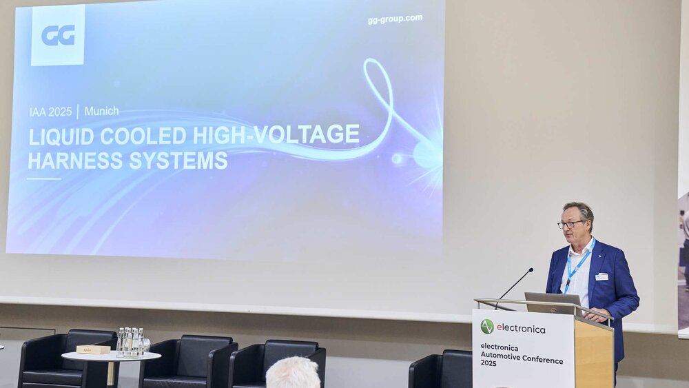 A man speaks at a podium next to a screen displaying the words LIQUID COOLED HIGH-VOLTAGE HARNESS SYSTEMS.