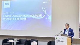A man speaks at a podium next to a screen displaying the words LIQUID COOLED HIGH-VOLTAGE HARNESS SYSTEMS.