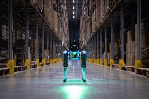  Four-legged robot stands in a high-bay warehouse between pallet racks.