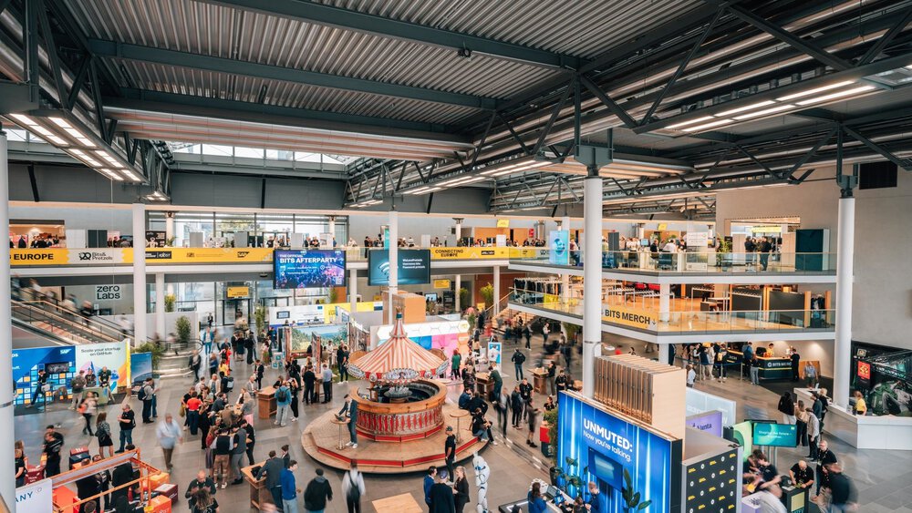 Der Blick über eine gut besuchte Messehalle mit mehreren Ebenen, Messeständen, Stützpfeilern und dunkelgrünen Stahlstreben an der Decke.